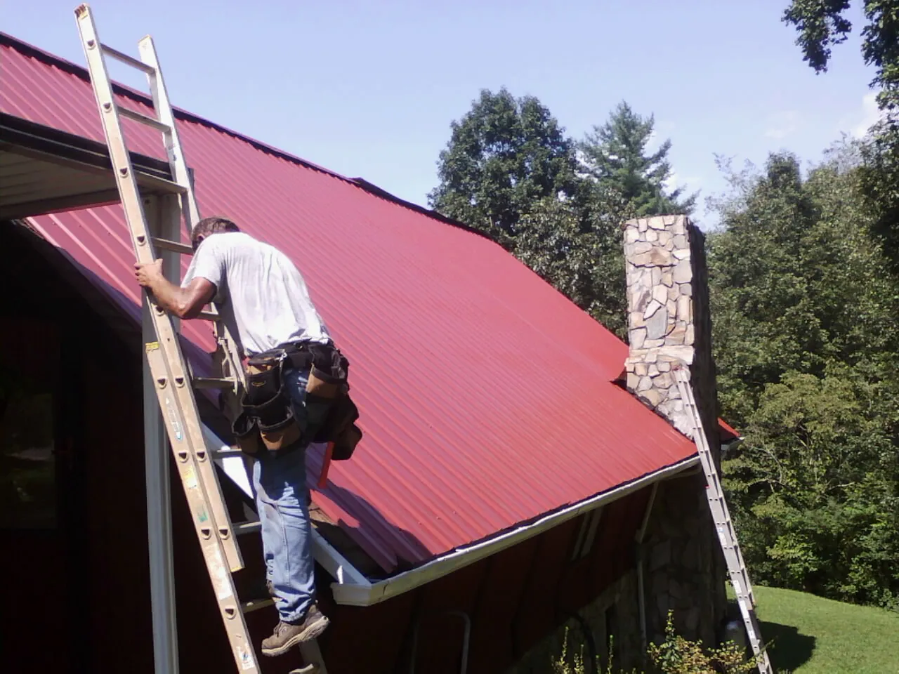 Roofer on a ladder beside a red metal roof and stone chimney — Soffit & Fascia Repair by Roofing Brothers in Lawnside